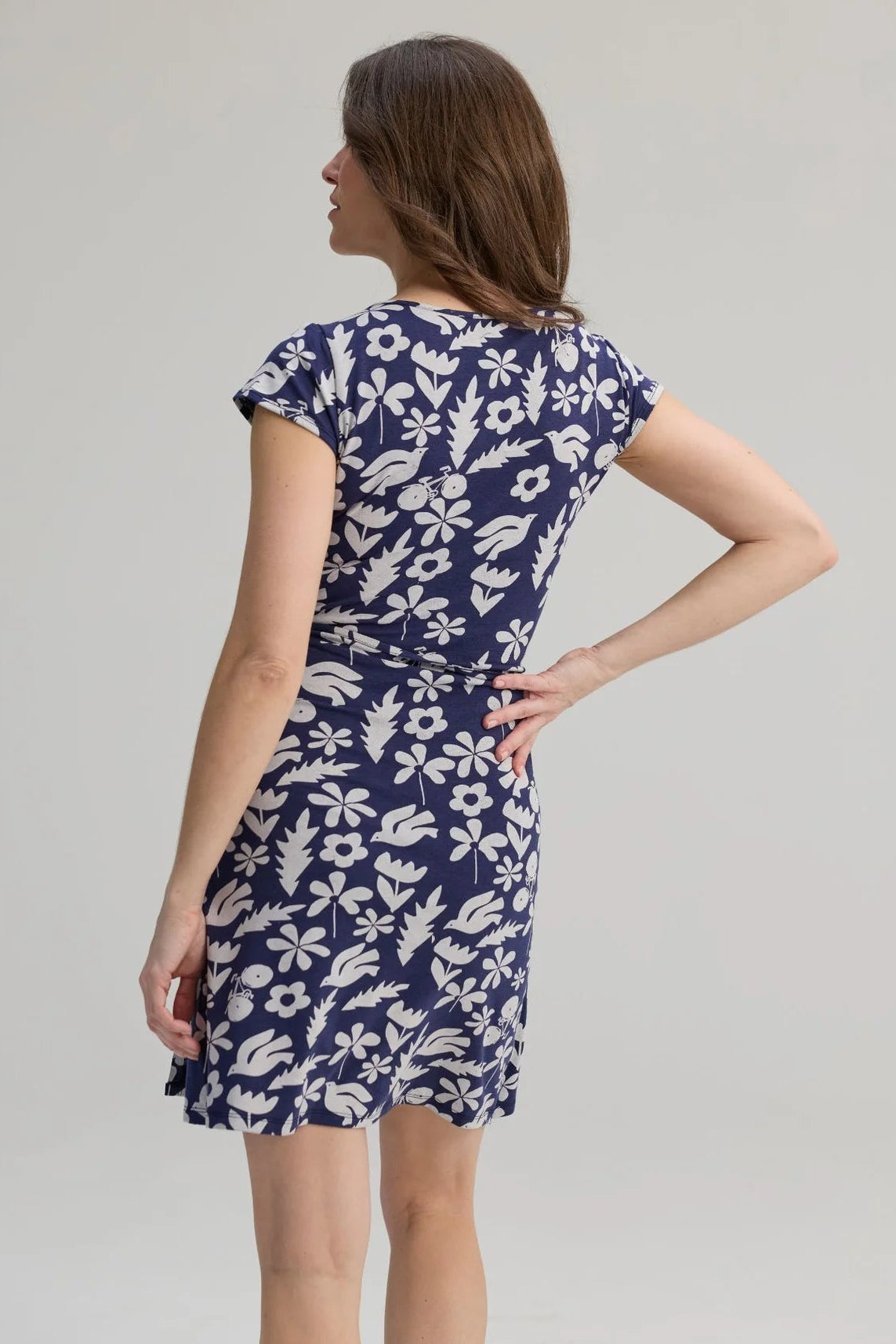 Back view of a woman wearing the Libellule Dress by Message Factory in Blue Print, a short sleeved wrap dress that falls to the knee. She is standing against a plain background. 