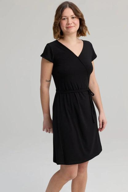 A woman wearing the Libellule Dress by Message Factory in Black, a short sleeved wrap dress that falls to the knee. She is standing against a plain background. 