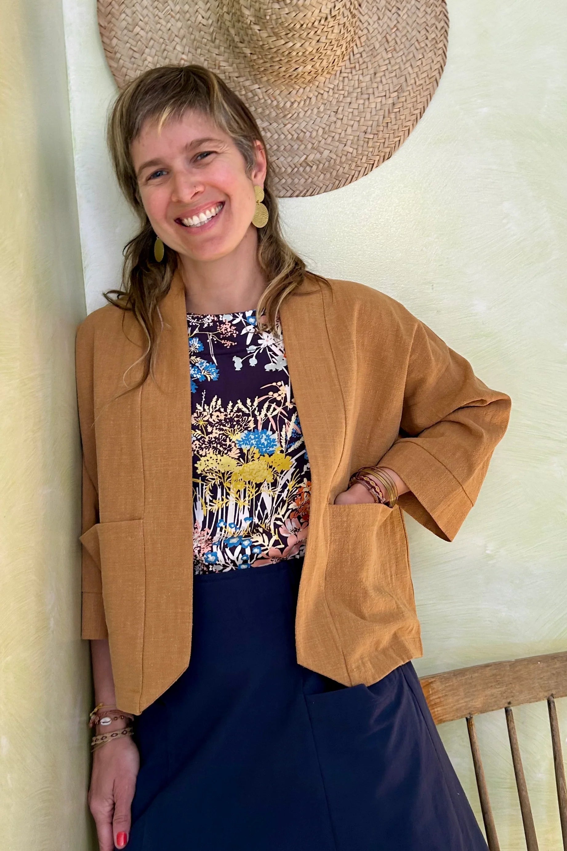 A woman wearing the Magnolia Jacket from Kazak in Turmeric, a kimono style jacket with 3/4 sleeves and large patch pockets. She is wearing it with a floral top navy and standing in a room with a wooden chair. 