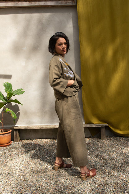 Side view of a woman wearing the Magnolia Screen-printed Jacket from Kazak in Olive with Dandelions, a short kimono style jacket with hand-screen printed dandelions, open front, 3/4-length sleeves, and big patch pockets. She is wearing it with matching pants and standing outside by a grey wall with a yellow curtain. 
