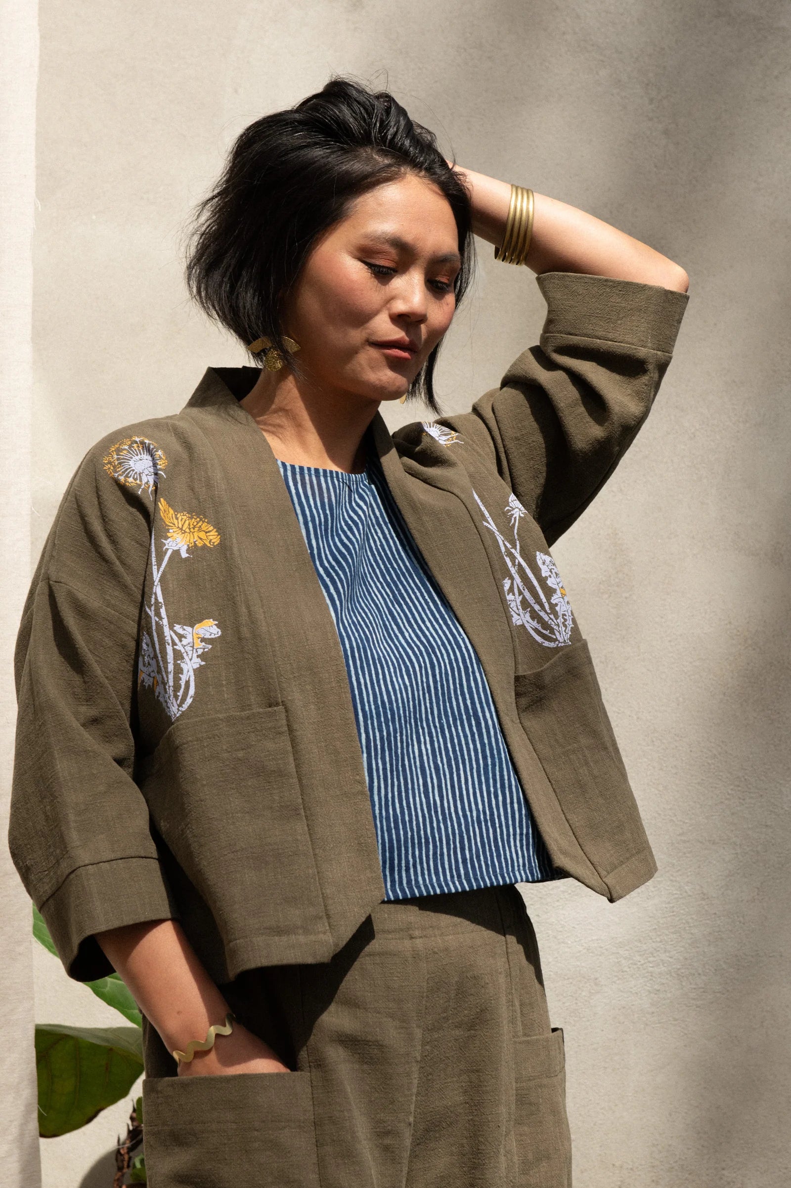 A woman wearing the Magnolia Screen-printed Jacket from Kazak in Olive with Dandelions, a short kimono style jacket with hand-screen printed dandelions, open front, 3/4-length sleeves, and big patch pockets. She is wearing it with matching pants and standing outside by a grey wall. 
