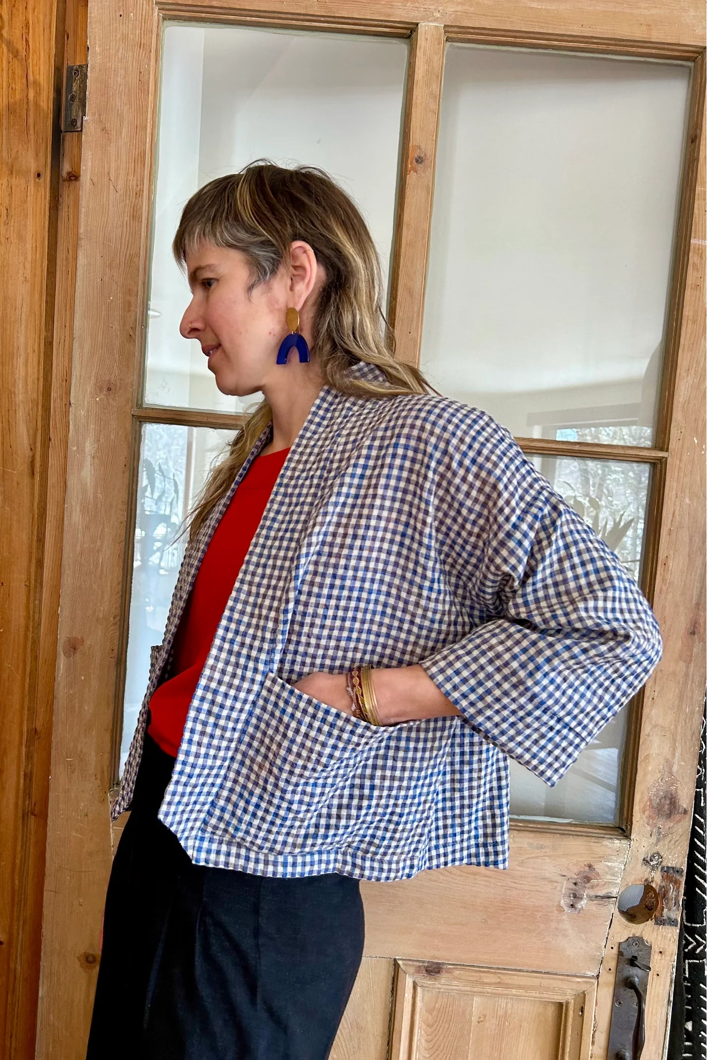 Side view of a woman wearing the Magnolia Jacket from Kazak in Sky Vichy/Gingham print, a kimono style jacket with 3/4 sleeves and large patch pockets. She is wearing it with a red top and navy skirt and standing in front of a door.
