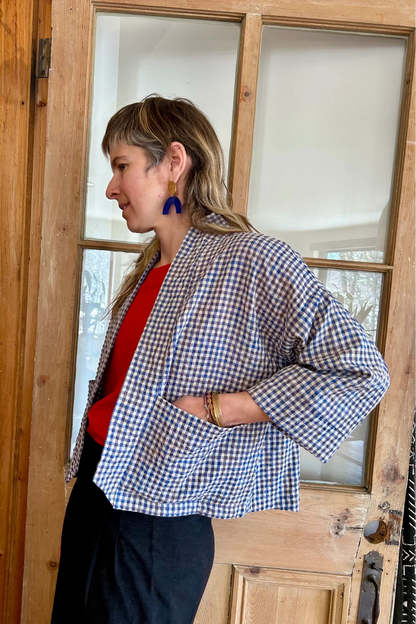 Side view of a woman wearing the Magnolia Jacket from Kazak in Sky Vichy/Gingham print, a kimono style jacket with 3/4 sleeves and large patch pockets. She is wearing it with a red top and navy skirt and standing in front of a door.