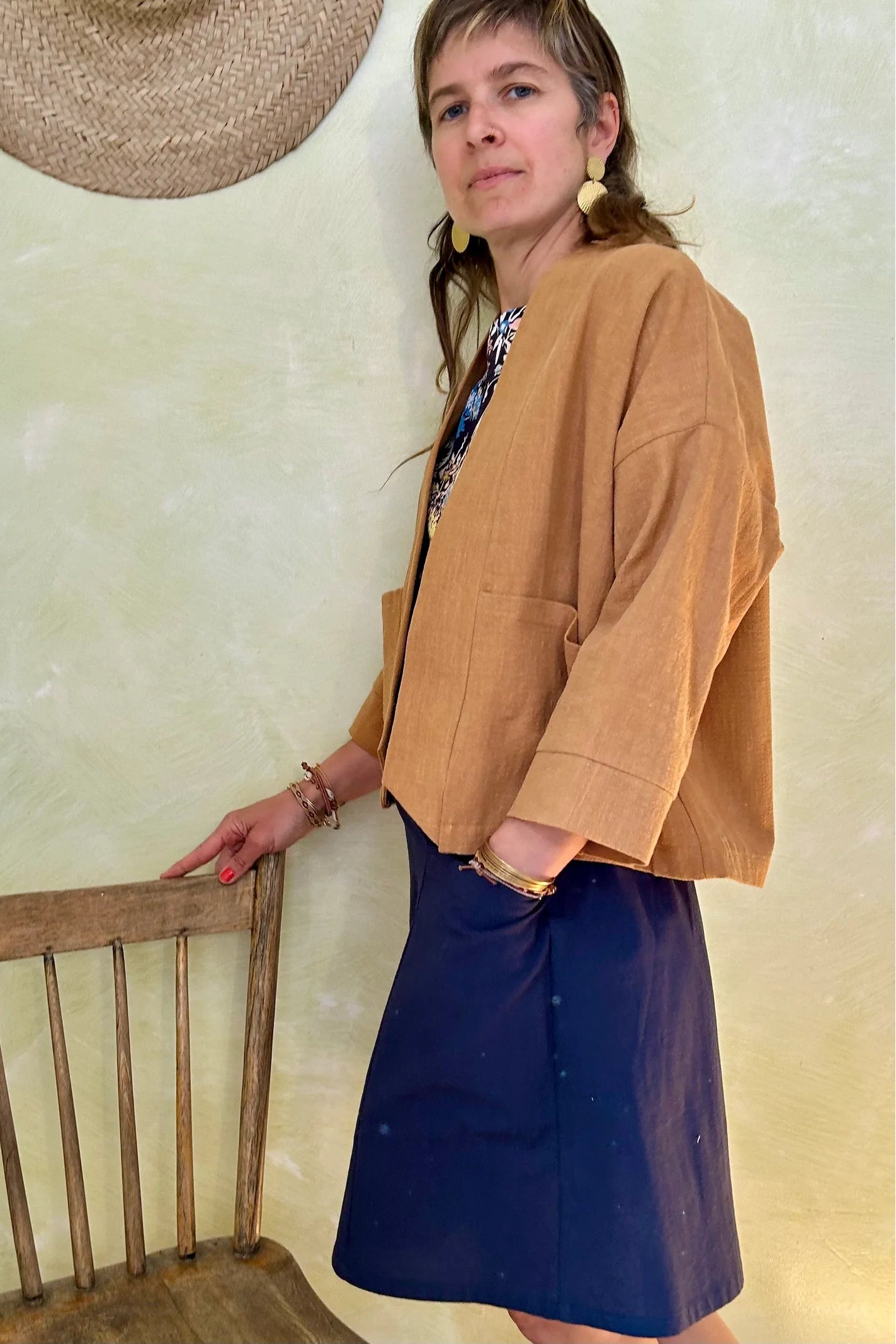 Side view of a woman wearing the Magnolia Jacket from Kazak in Turmeric, a kimono style jacket with 3/4 sleeves and large patch pockets. She is wearing it with a floral top navy and standing in a room with a wooden chair. 