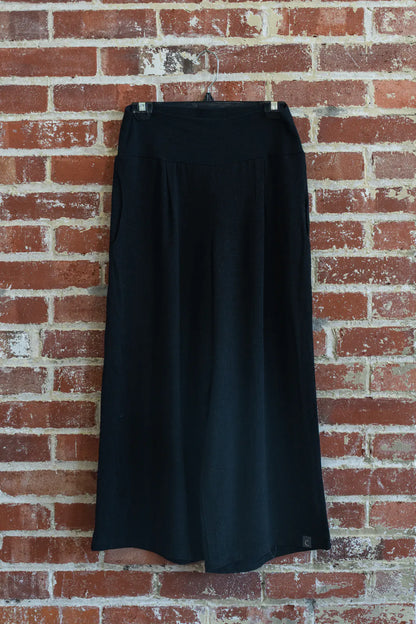 The Verano Pants by Marie C in Black are shown hanging in front of a brick wall. 