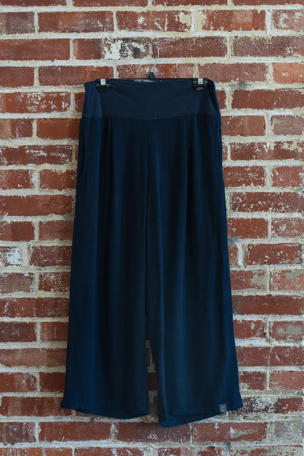 The Verano Pants by Marie C in Navy are shown hanging in front of a brick wall. 