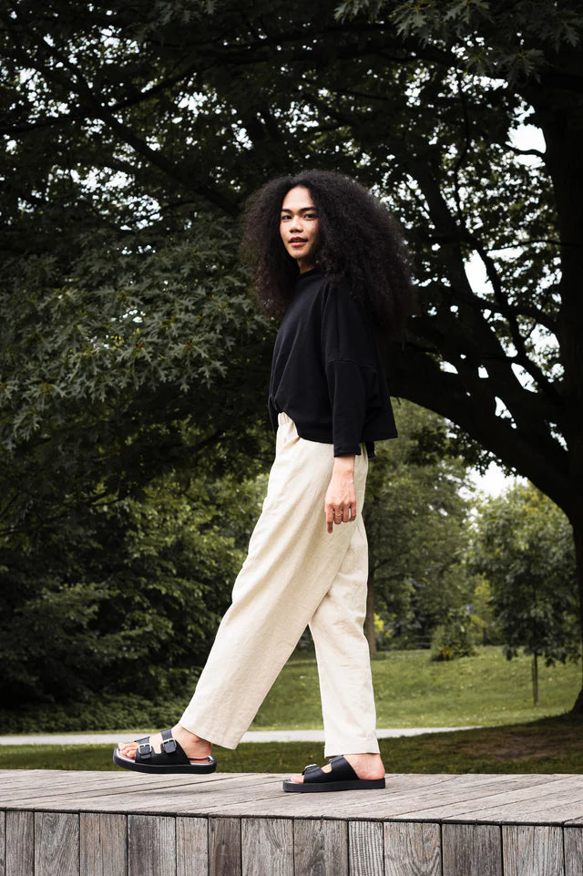 Woman wearing the Bogota Top by MAS in Pepper, an oversized top with a high neck, exaggerated dropped shoulders, and long sleeves, in ribbed fabric. She is wearing it with cream pants and standing outside on a wooden platform. 