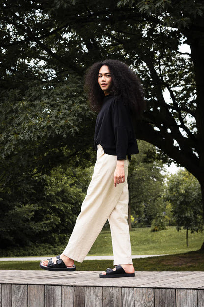 Woman wearing the Bogota Top by MAS in Pepper, an oversized top with a high neck, exaggerated dropped shoulders, and long sleeves, in ribbed fabric. She is wearing it with cream pants and standing outside on a wooden platform. 