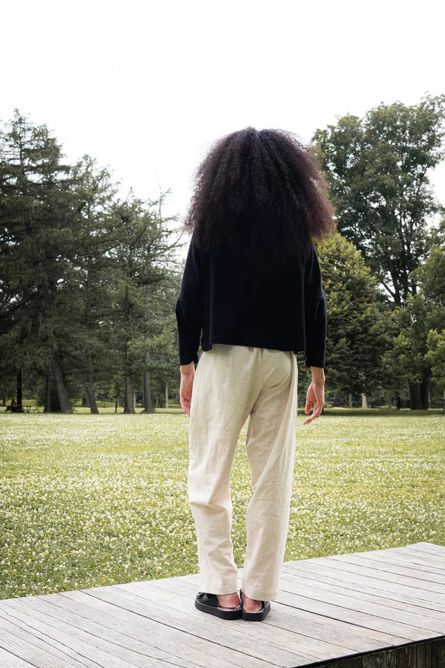 Woman wearing the Bogota Top by MAS in Pepper, an oversized top with a high neck, exaggerated dropped shoulders, and long sleeves, in ribbed fabric. She is wearing it with cream pants and standing outside on a wooden platform