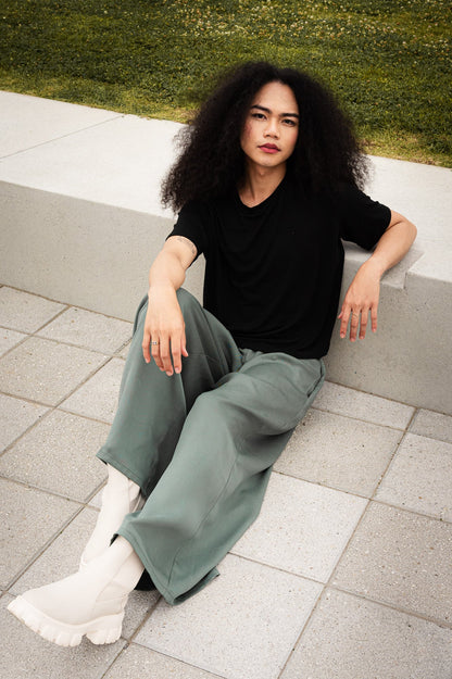 A woman wearing the Santiago T-Shirt by MAS in Pepper, a classic tee witha slightly cropped and boxy fit. She is wearing it with green pants and sitting outside on a paved structure. 