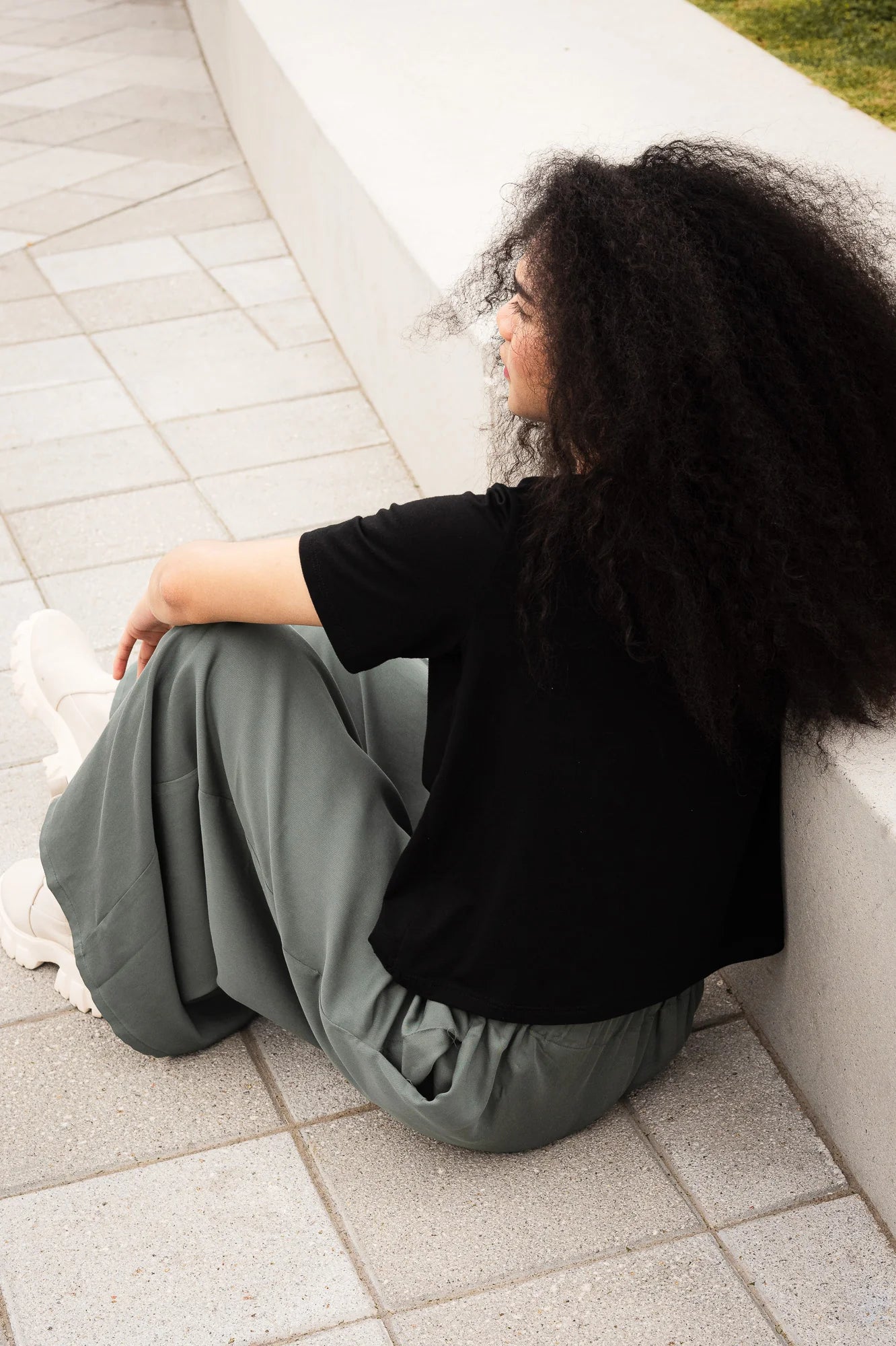 Back view of a woman wearing the Santiago T-Shirt by MAS in Pepper, a classic tee witha slightly cropped and boxy fit. She is wearing it with green pants and sitting outside on a paved structure. 
