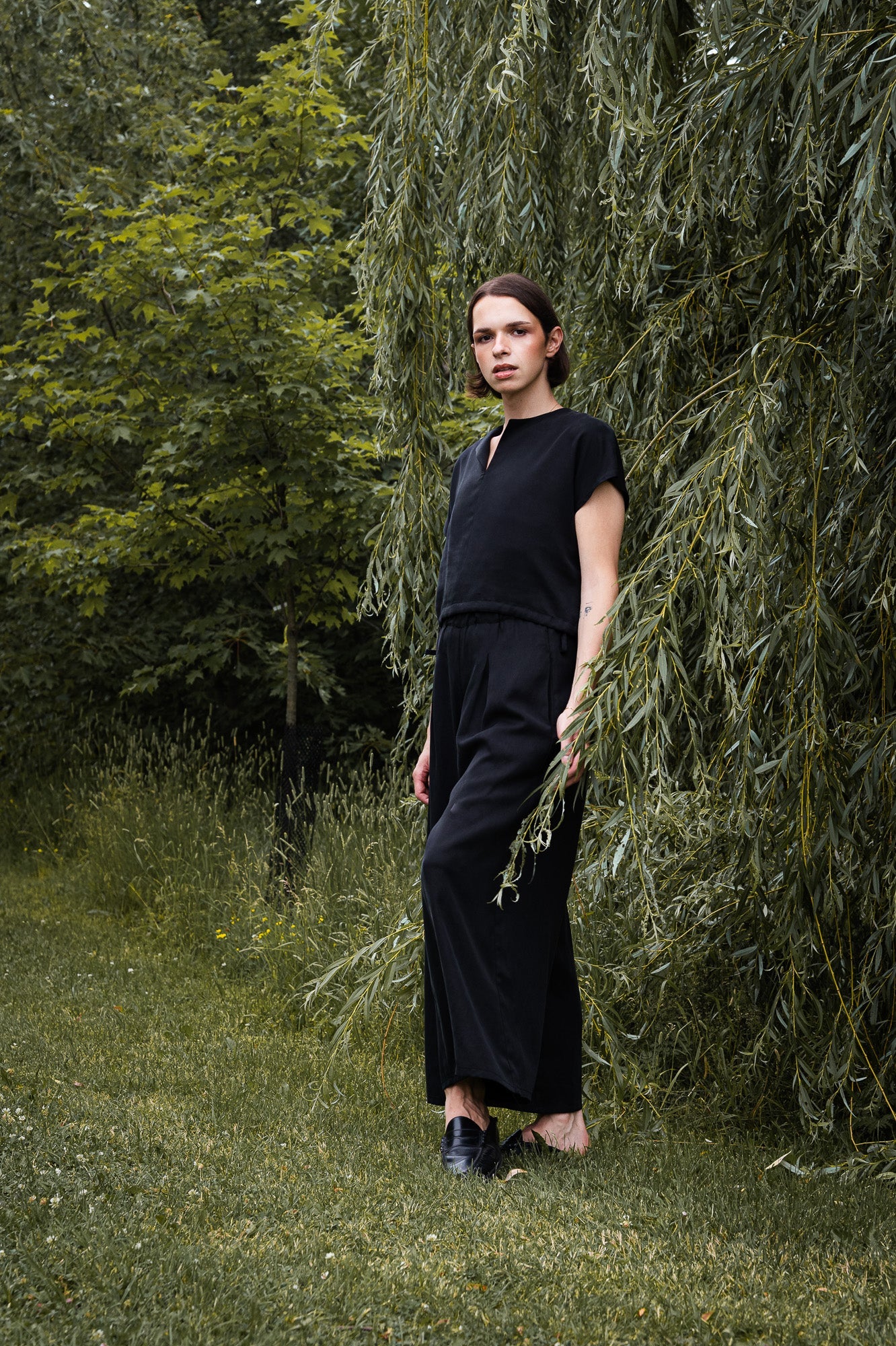 A woman wearing the Siena Pants by MAS in Pepper, wide legged trousers with an elastic waist. She is wearing them with a black top and standing outdoors among trees.
