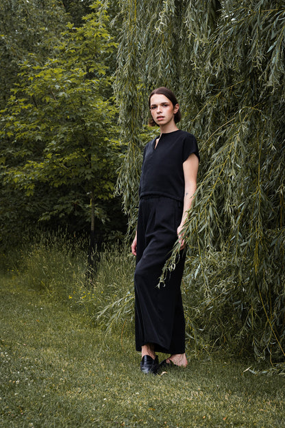A woman wearing the Siena Pants by MAS in Pepper, wide legged trousers with an elastic waist. She is wearing them with a black top and standing outdoors among trees.
