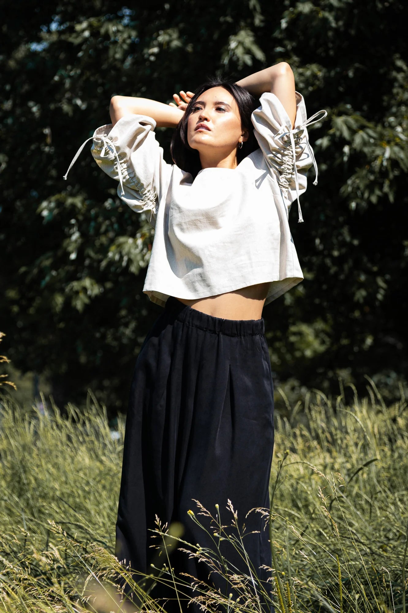 A woman wearing the Siena Pants by MAS in Pepper, wide legged trousers with an elastic waist. She is wearing them with a white top and standing outdoors. 