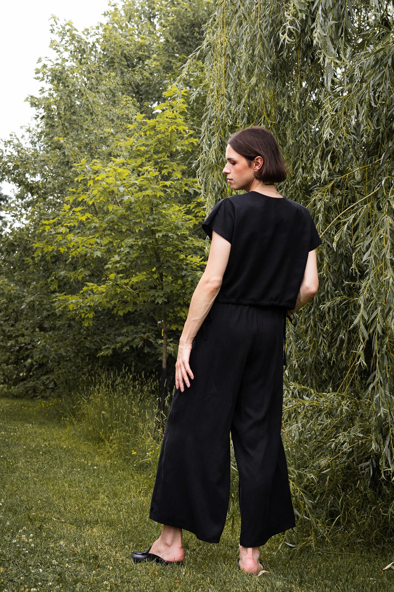 Back view of a woman wearing the Siena Pants by MAS in Pepper, wide legged trousers with an elastic waist. She is wearing them with a black top and standing outdoors among trees.