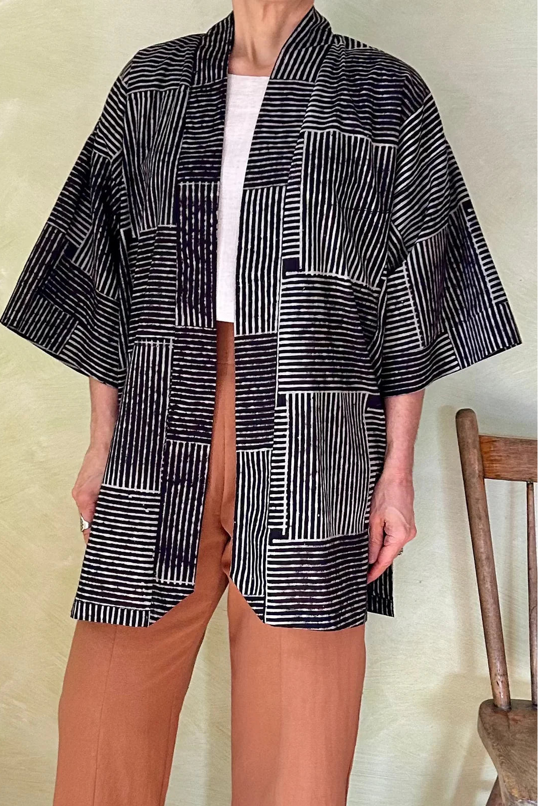Neck down view of a woman wearing the Mirano Jacket by Kazak in Oris Slate, a cross hatch pattern. It is a  mid-thigh length open kimono jacket with 3/4 sleeves. She is wearing it with the Oxalis pants in Ginger and standing in a room by a wooden chair. 