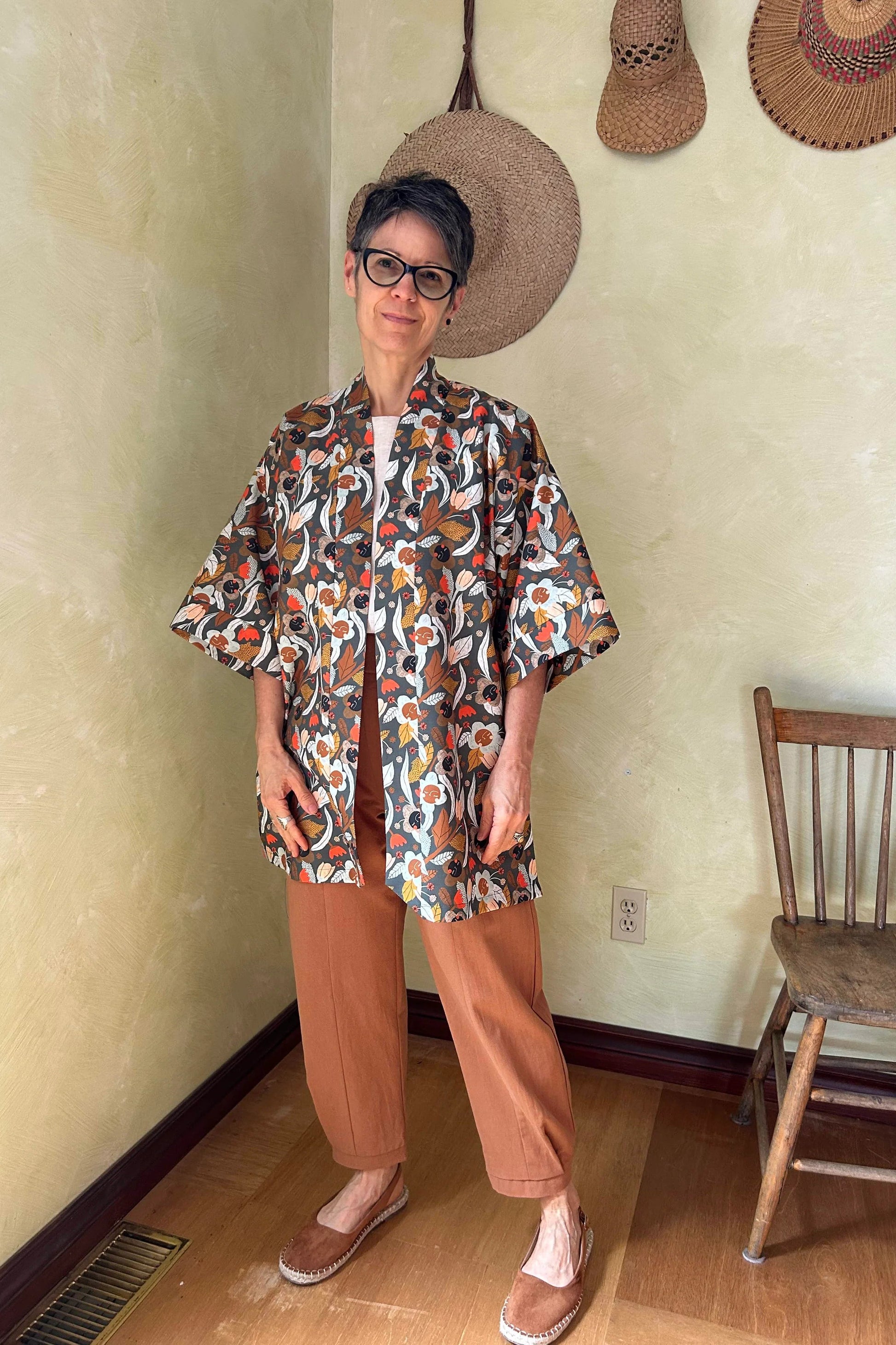 A woman wearing the Mirano Jacket by Kazak in Olive Floral, a mid-thigh length open kimono jacket with 3/4 sleeves. She is wearing it with the Oxalis pants in Ginger and standing in a room by a wooden chair. 