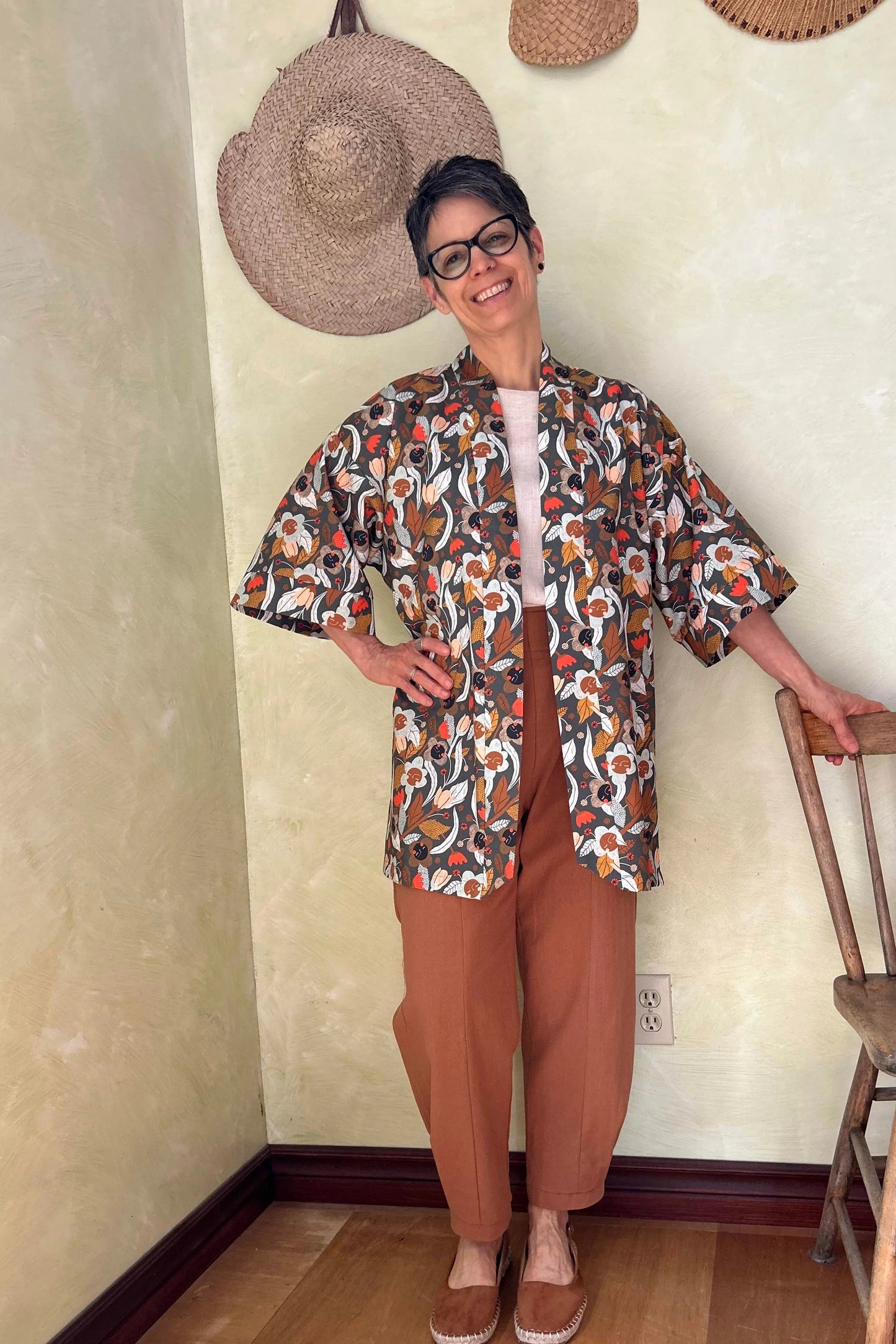 A woman wearing the Mirano Jacket by Kazak in Olive Floral, a mid-thigh length open kimono jacket with 3/4 sleeves. She is wearing it with the Oxalis pants in Ginger and standing in a room by a wooden chair. 