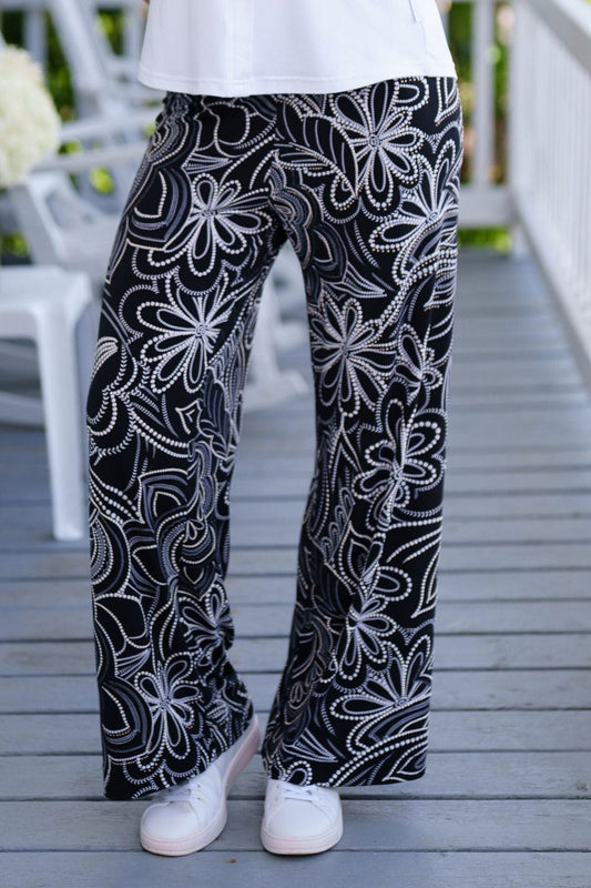 Waist down view of A woman wearing the Coco Palazzo Pants by Rien ne se Perd in Black and White floral, with a wide elastic waistband and wide legs. She is wearing them with a white top and standing on a wooden porch.