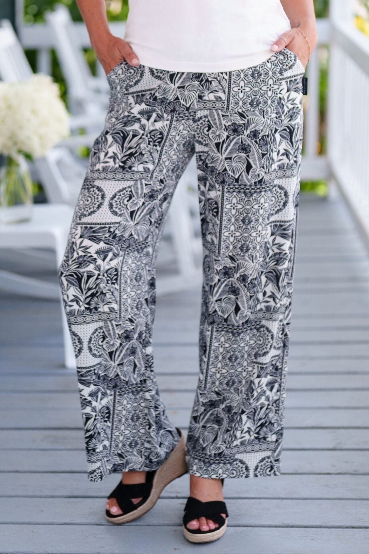 Waist down view of a woman wearing the Mango Pants from Rien ne se Perd in Black/Ivory tropical print, palazzo pants with an elastic waist and pockets. She is wearing them with a white top and standing on a porch. 