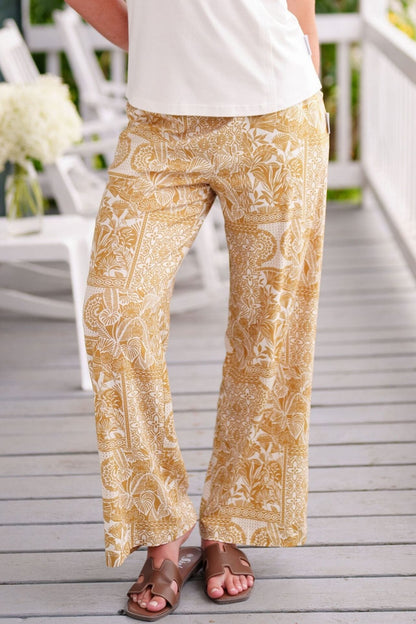 Waist down view of a woman wearing the Mango Pants from Rien ne se Perd in Ocre/Ivory tropical print, palazzo pants with an elastic waist and pockets. She is wearing them with a white top and standing on a porch. 