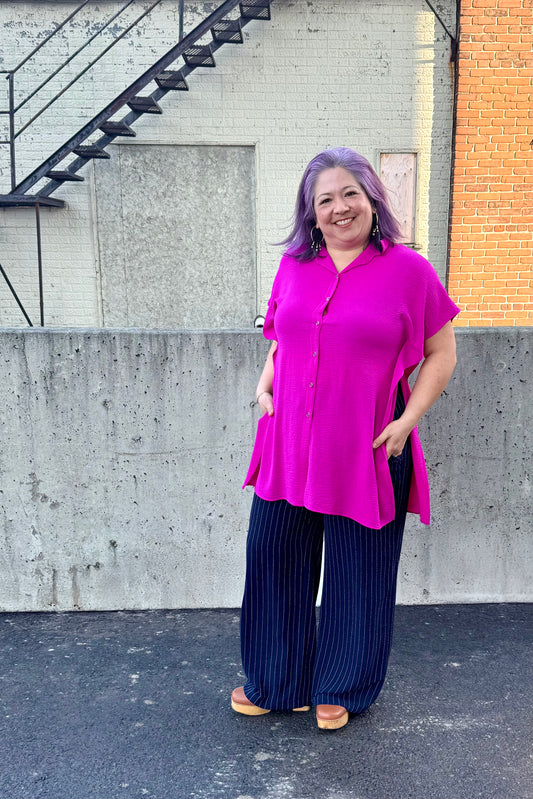 A woman wearing the Geneva Top by Pure Essence in Magenta, a long and loose button front shirt with short sleeves, a classic collar, and high side slits. She is wearing it with the Gabby Pants in navy/white pinstripe and standing outdoors against a concrete wall. 