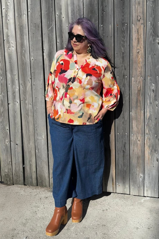 A woman wearing the Grace Blouse by Pure Essence in Coral Meadow, with a v-placket neck with buttons and 7/8 gathered sleeves. She is wearing it with denim pants and  standing against a wooden fence.