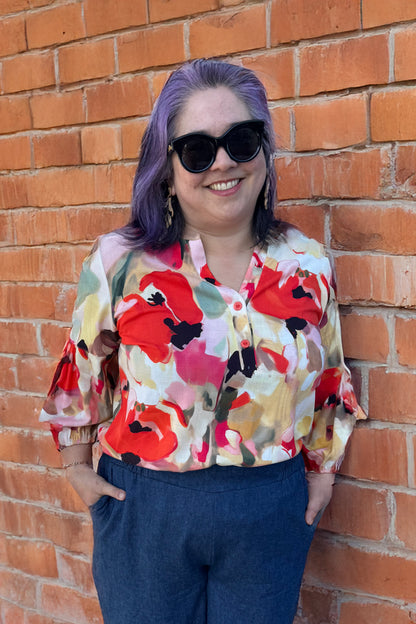A woman wearing the Grace Blouse by Pure Essence in Coral Meadow, with a v-placket neck with buttons and 7/8 gathered sleeves. She is wearing it with denim pants and by a brick wall. 