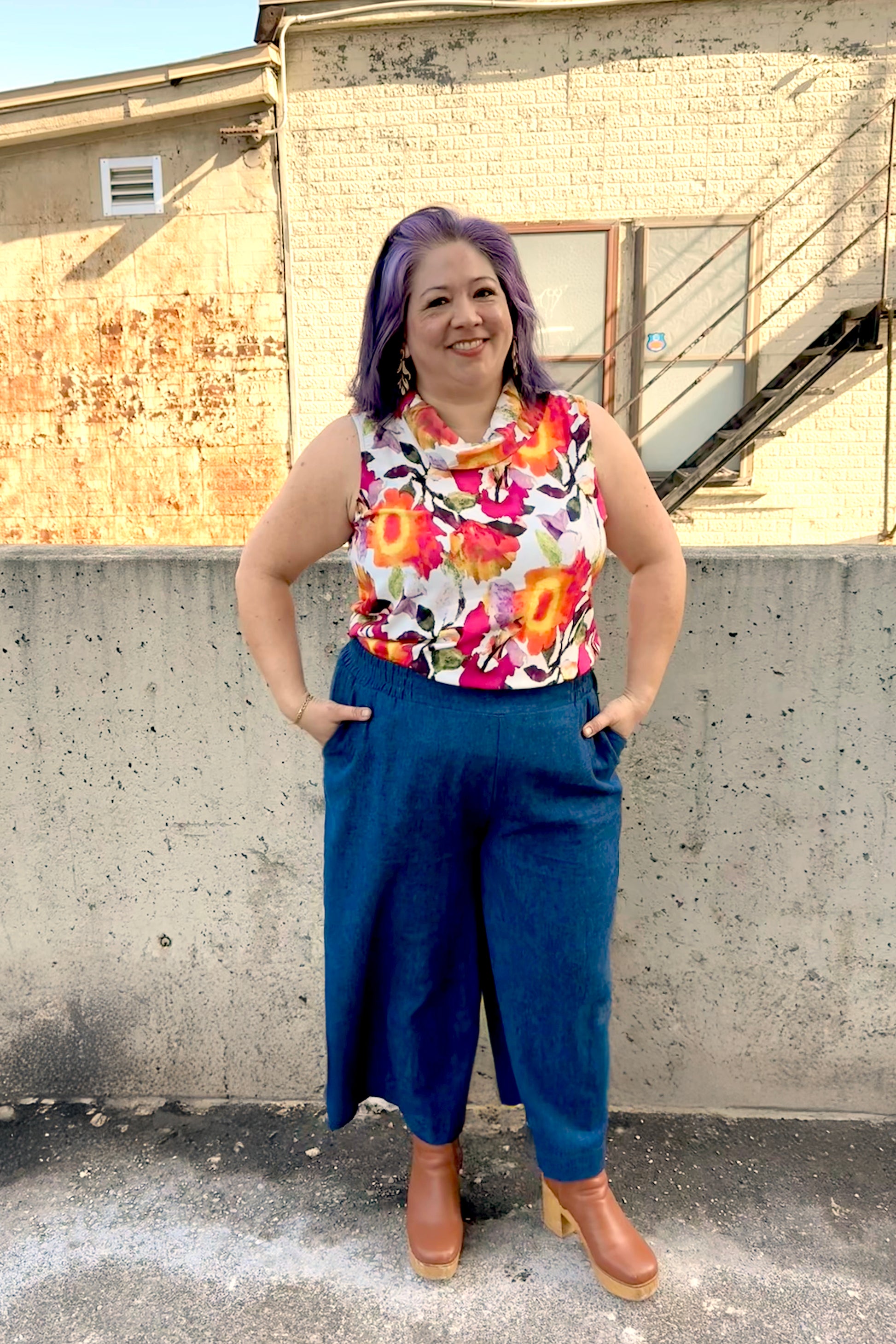 A woman wearing the Gemma top from Pure Essence in Wild Bloom, a floral sleeveless top with a cowl neck. She is wearing it with the Mateo pants from Melow in Denim and standing in front of a brick building.