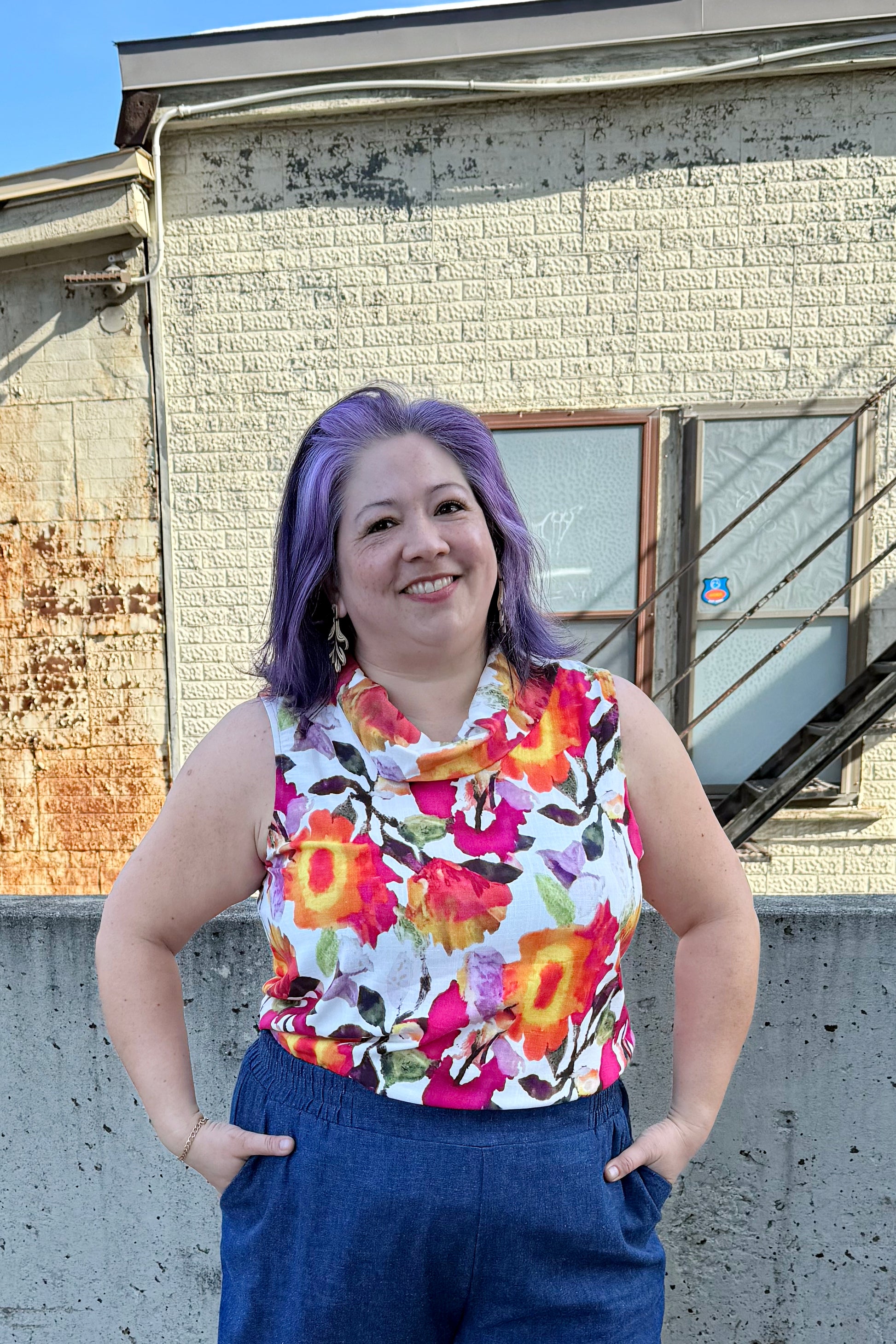 A woman wearing the Gemma top from Pure Essence in Wild Bloom, a floral sleeveless top with a cowl neck. She is wearing it with the Mateo pants from Melow in Denim and standing in front of a brick building.