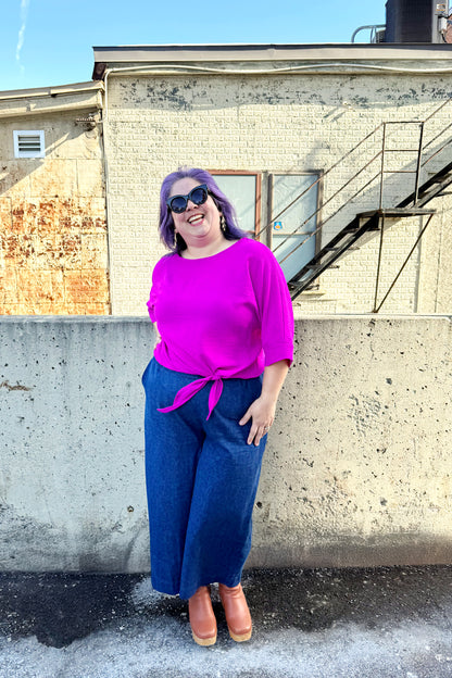 A woman wearing the Ginger Top from Pure Essence in Magenta, with a wide neck, blousy fit, 3/4 sleeves, and tie detail at the waist. She is wearing it with the Mateo Pants by Melow in Denim and standing outside in front of a brick building. 