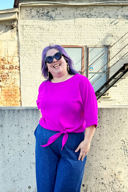 A woman wearing the Ginger Top from Pure Essence in Magenta, with a wide neck, blousy fit, 3/4 sleeves, and tie detail at the waist. She is wearing it with the Mateo Pants by Melow in Denim and standing outside in front of a brick building. 