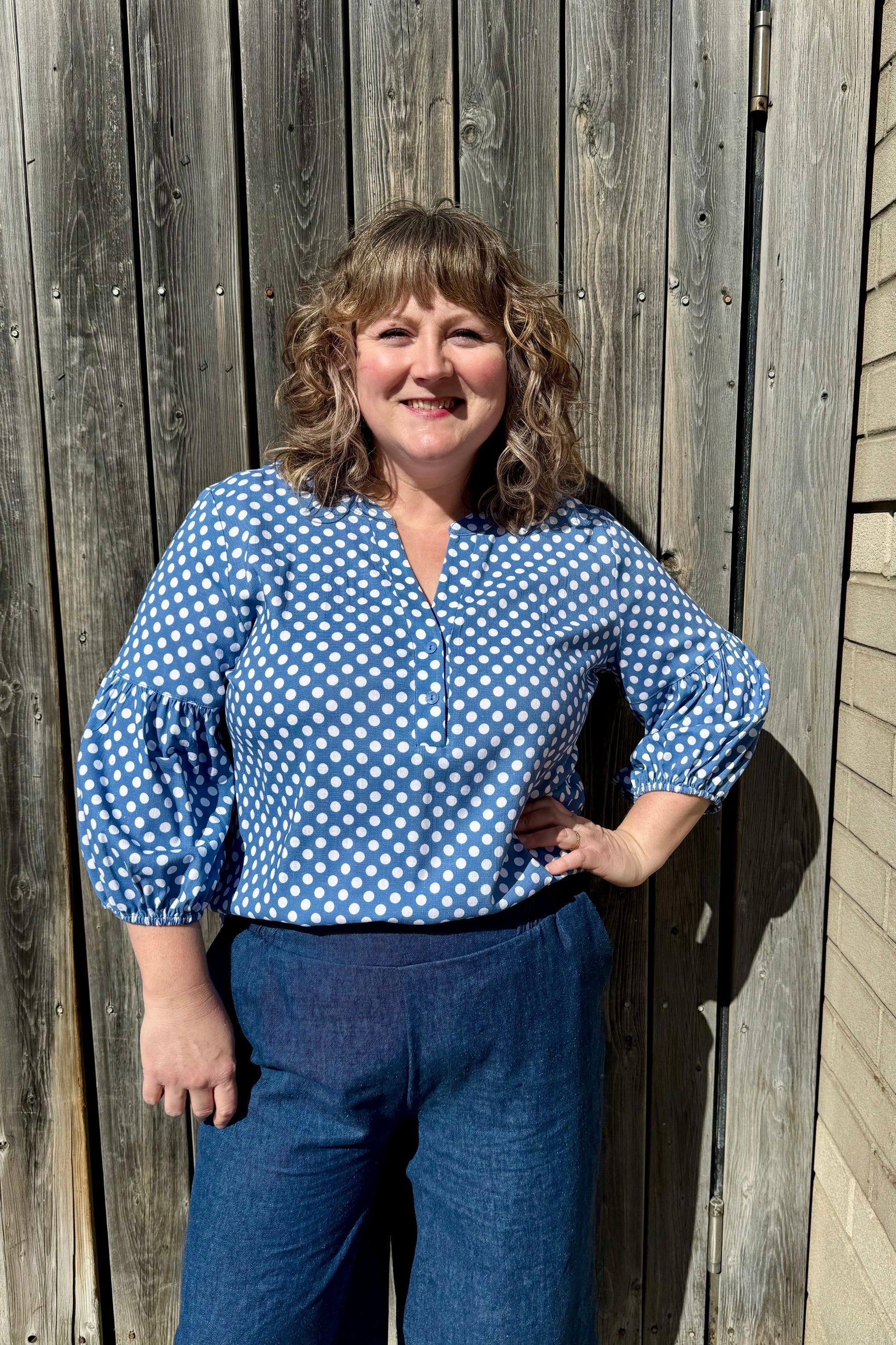 A woman wearing the Grace Blouse by Pure Essence in Chambray Dot, with a v-placket neck with buttons and 7/8 gathered sleeves. She is wearing it with denim pants and standing against a wooden fence 