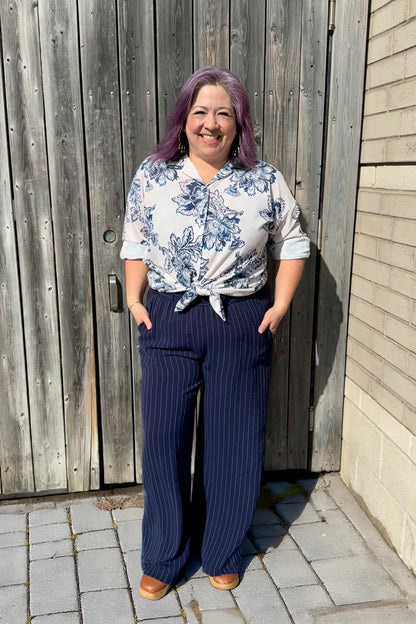 A woman wearing the Garnet Shirt by Pure Essence in Blue Stone floral print,  a classic button front shirt with dropped shoulders.  She is wearing with the Gabby Pants and standing in front of wooden fence.