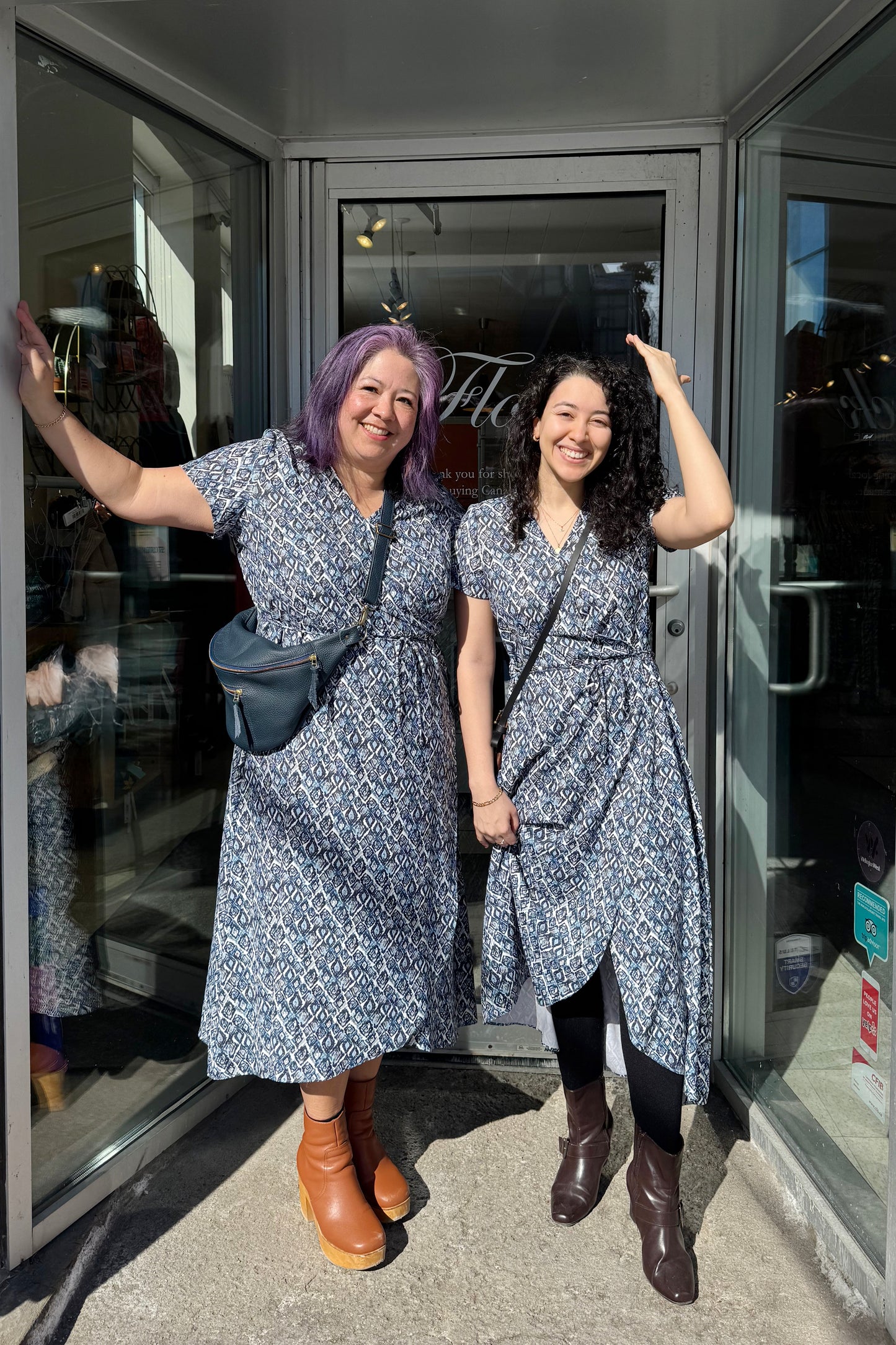 Two women in matching Galea Dresses by Pure Essence in Blue Lattice print. This faux-wrap dress has  short sleeves, a gathered waist with a tie and a midi-length tulip hem. They are standing outside the Flock entrance.