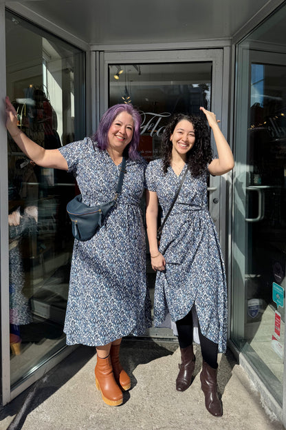 Two women in matching Galea Dresses by Pure Essence in Blue Lattice print. This faux-wrap dress has  short sleeves, a gathered waist with a tie and a midi-length tulip hem. They are standing outside the Flock entrance.