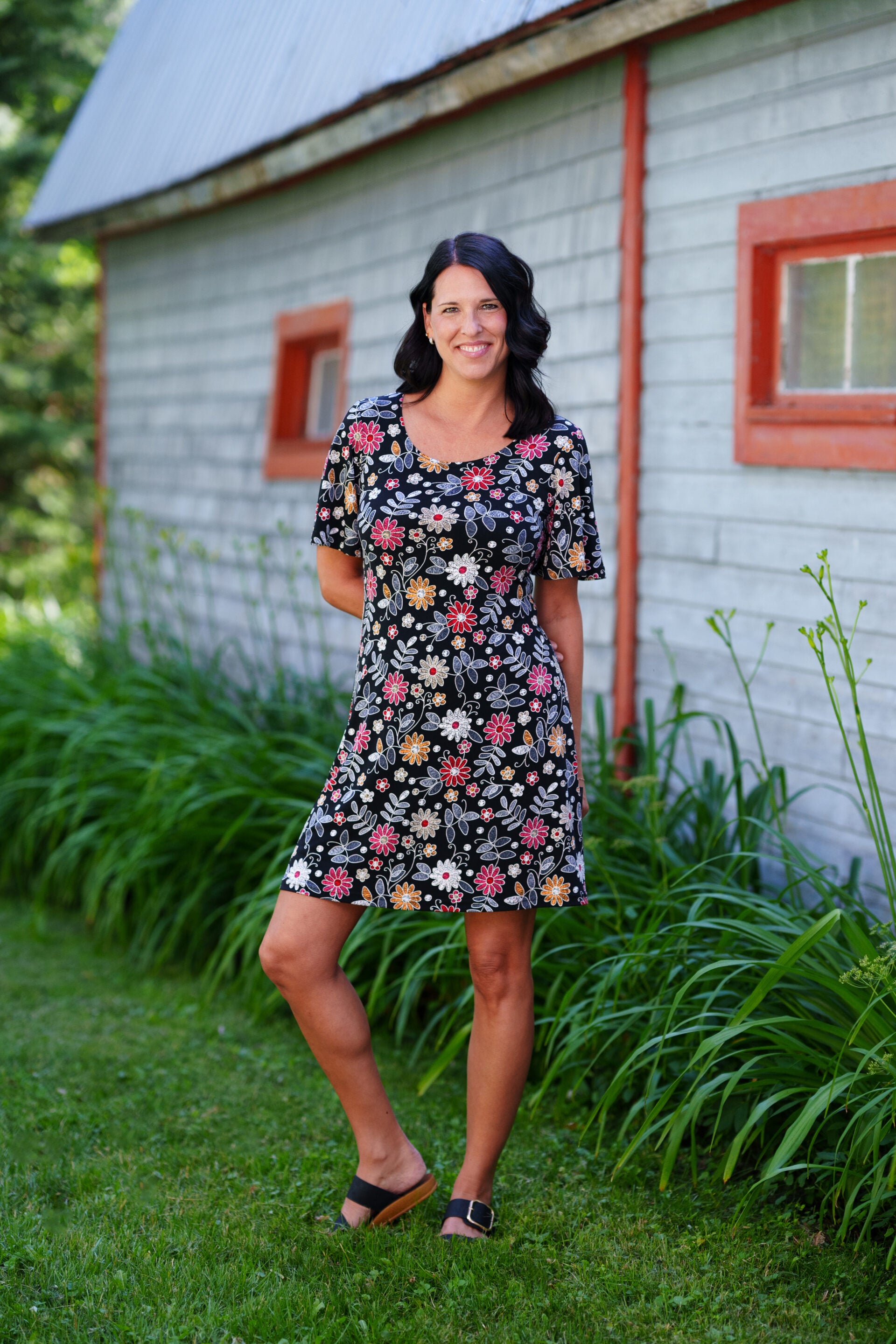 A woman wearing the Grenadine Dress by Rien ne se Perd in Floral print,  a short A-line dress with bell sleeves and a scoop neck. She is  standing in front of a rustic building with red windows.
