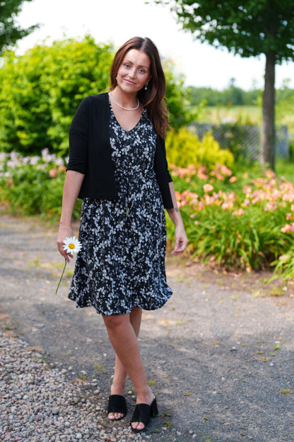 A woman wearing the Salvador Bolero in Black from Rien ne se Perd, featuring 3/4 sleeves and an angled hem, over the Mambo Dress. She is standing outside in a garden. 