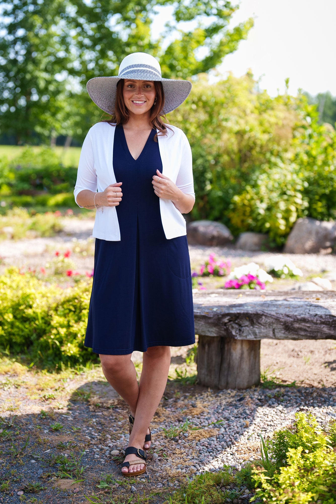 A woman wearing the Salvador Bolero in White from Rien ne se Perd, featuring 3/4 sleeves and an angled hem, over the Samba Dress in Navy. She is standing in a garden. 