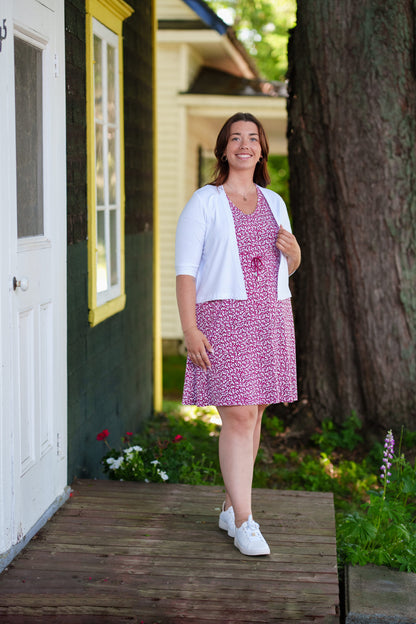 A woman wearing the Sangria Dress by Rien ne se Perd in Raspberry floral print, a short dress with a V-neck, an adjustable drawstring ribbon under the bust, and short sleeves. She is wearing it with a white bolero and standing on a door step with a tree behind her. 