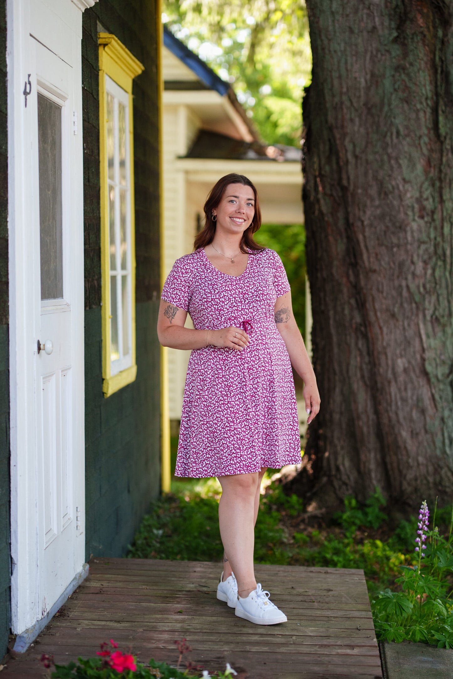 A woman wearing the Sangria Dress by Rien ne se Perd in Raspberry floral print, a short dress with a V-neck, an adjustable drawstring ribbon under the bust, and short sleeves. She is standing on a door step with a tree behind her. 