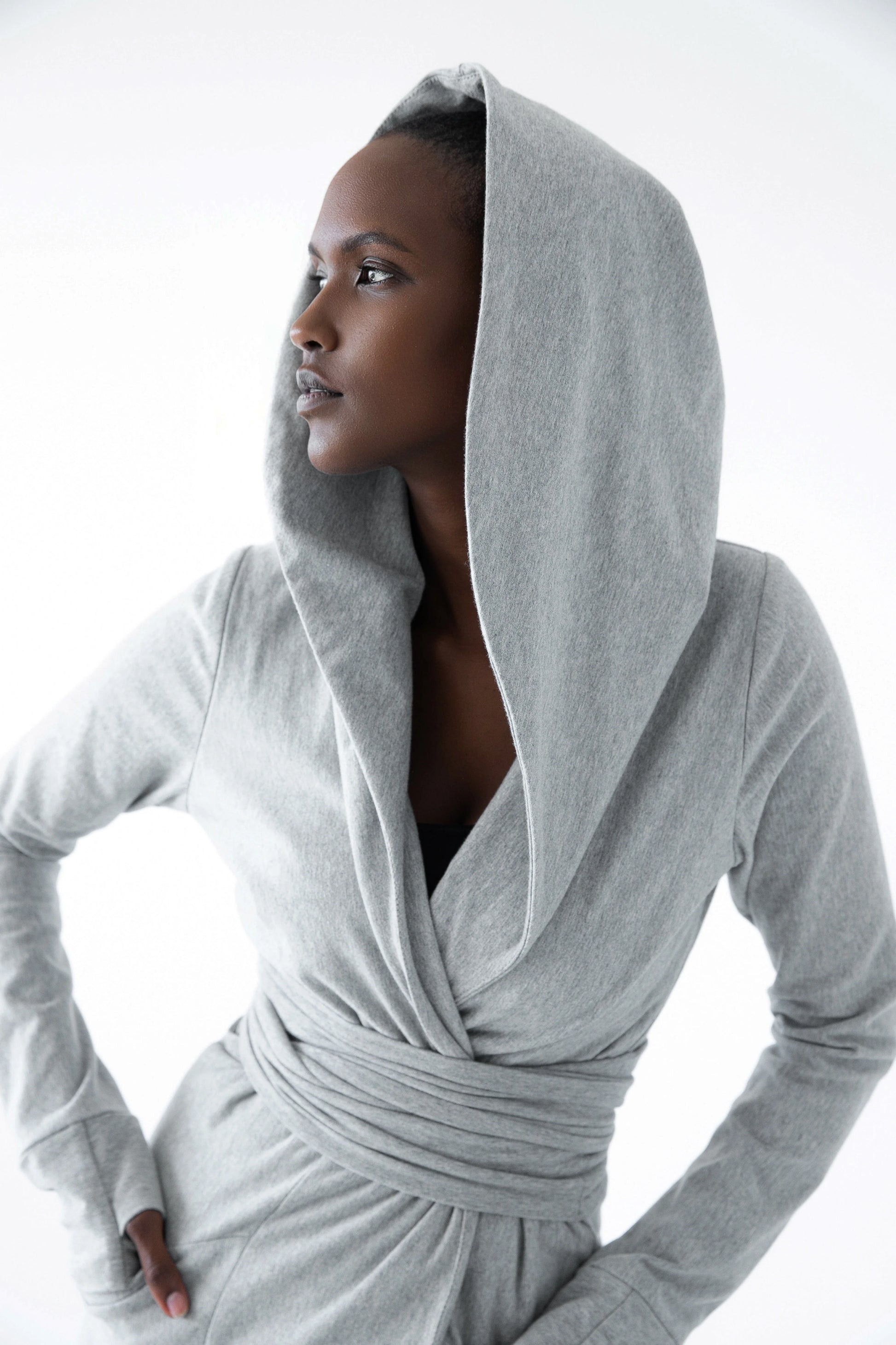 A woman wearing the Signature Cotton Hooded Wrap by Stacey Martin in Stone a hooded, corset waist sweatshirt with a tie belt. She is standing against a white background. 