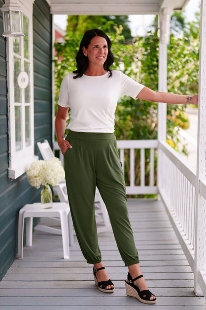 A woman wearing the Mai Tai T-shirt from Rien ne se Perd in Ivory, with coconut buttons at the shoulder, seam accents front and back, and a rounded hi-low hemline. She is wearing it with green pants and standing on a wooden porch. 