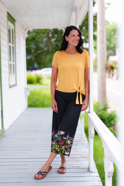 A woman wearing the Amaretto 7/8 Gaucho Pants from Rien ne se Perd in Black with a floral print hem. She is wearing them with a yellow top and standing on a porch. 