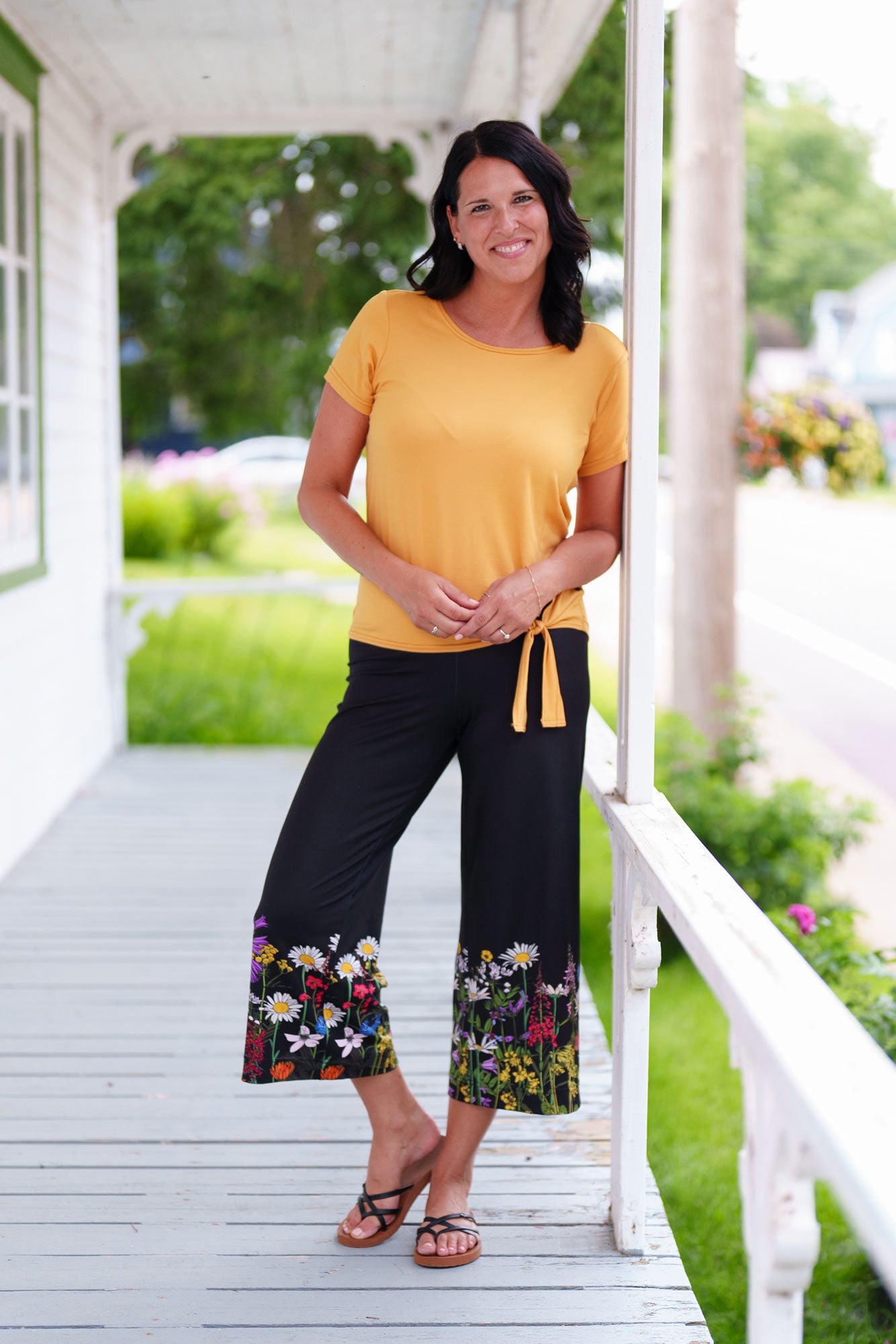 A woman wearing the Amaretto 7/8 Gaucho Pants from Rien ne se Perd in Black with a floral print hem. She is wearing them with a yellow top and standing on a porch. 