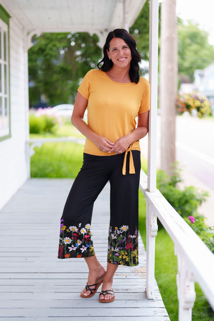 A woman wearing the Amaretto 7/8 Gaucho Pants from Rien ne se Perd in Black with a floral print hem. She is wearing them with a yellow top and standing on a porch. 