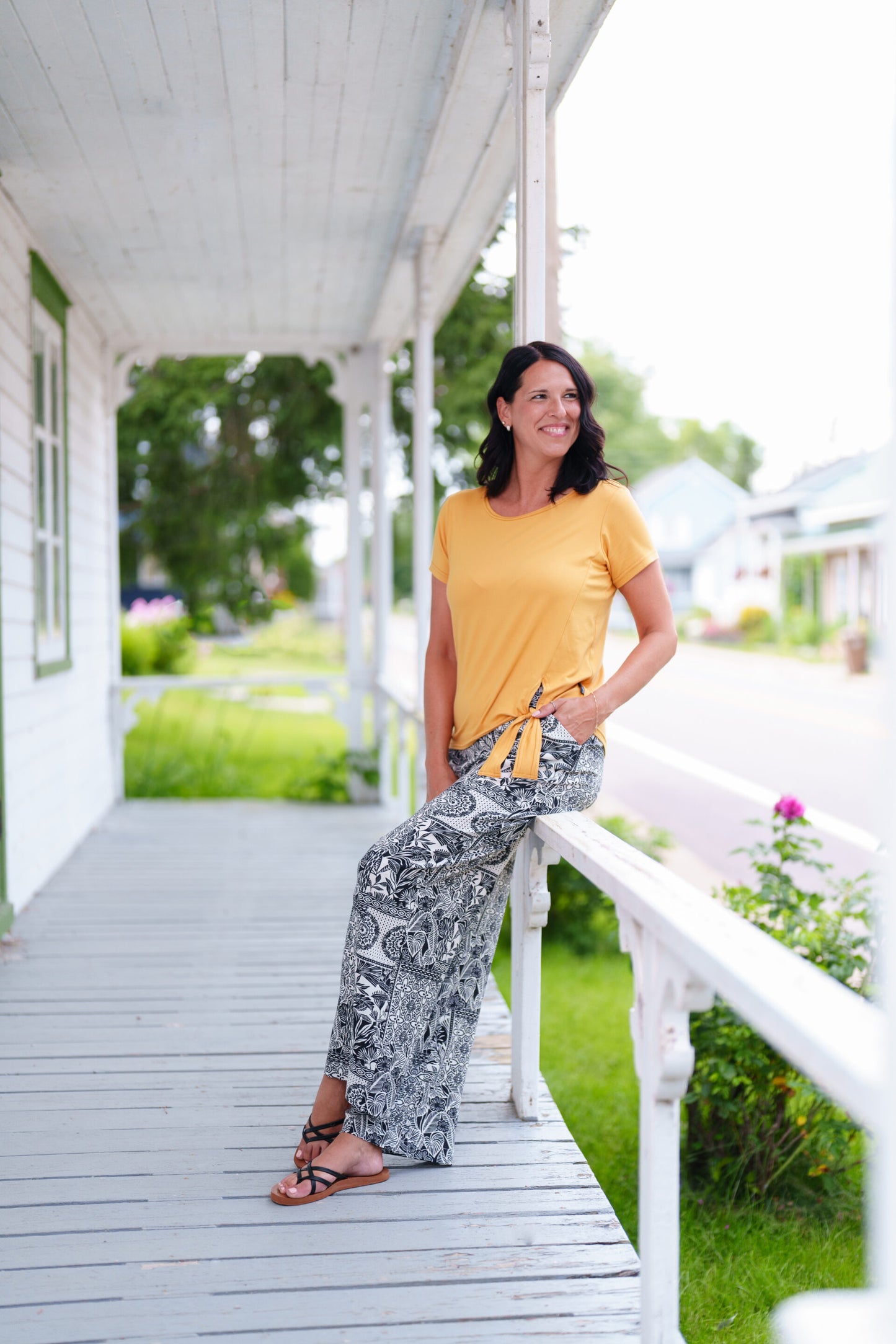 A woman wearing the Mango Pants from Rien ne se Perd in Black/Ivory tropical print, palazzo pants with an elastic waist and pockets. She is wearing them with ta yellow top and sitting on a porch railing. 