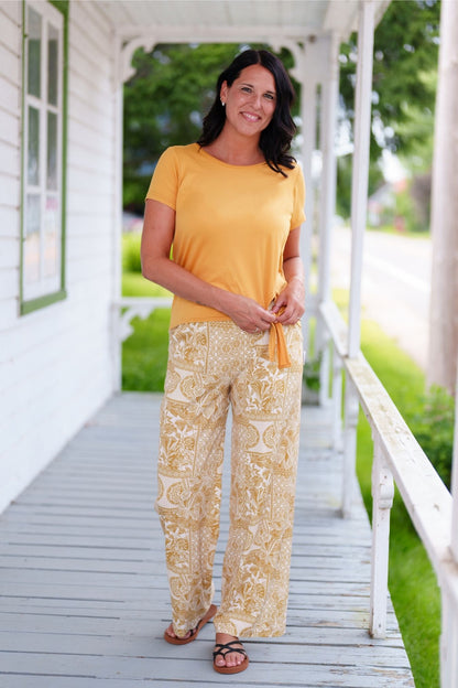 A woman wearing the Mango Pants from Rien ne se Perd in Ocre/Ivory tropical print, palazzo pants with an elastic waist and pockets. She is wearing them with a yellow top and standing on a porch. 