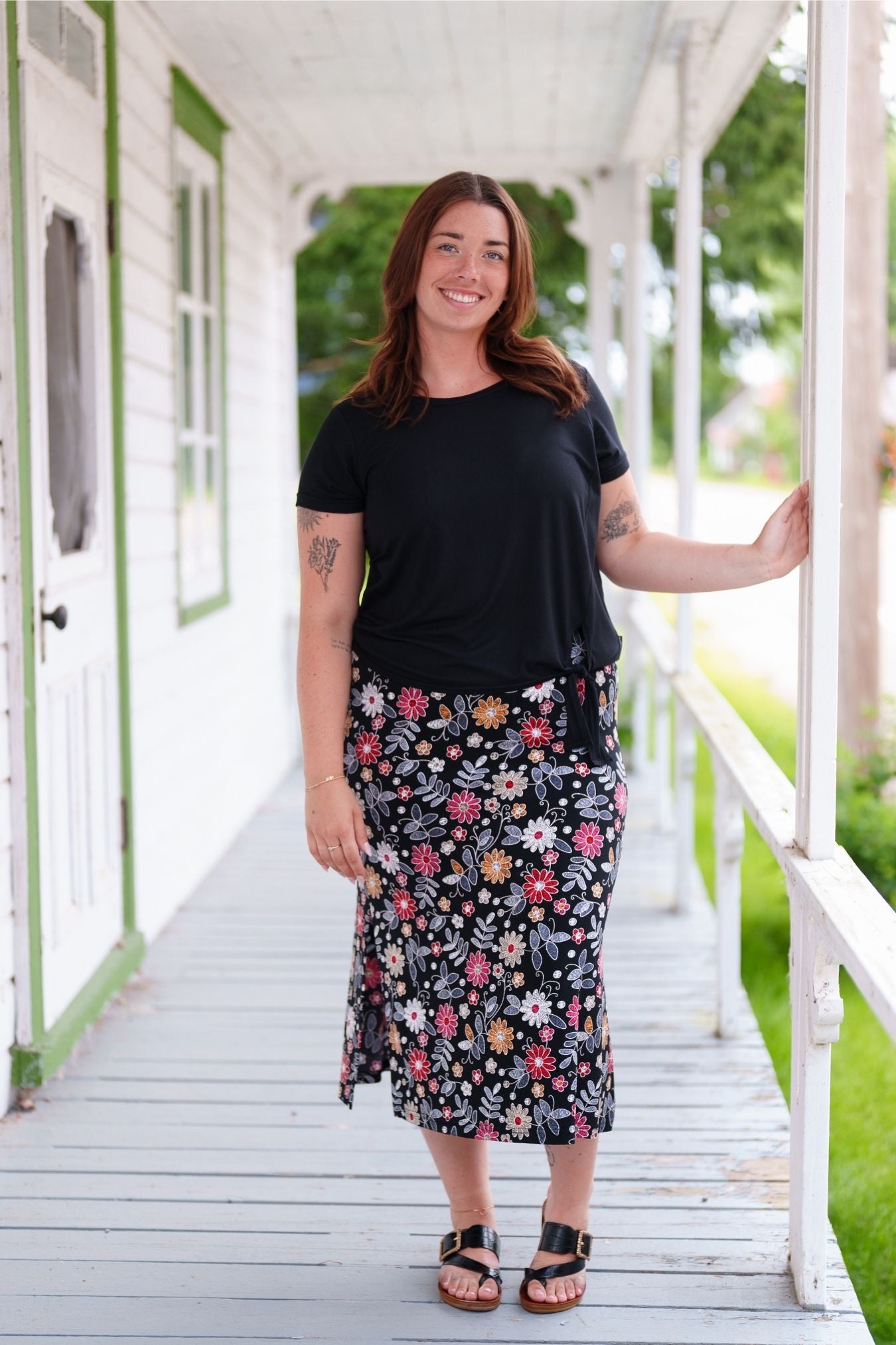 A woman wearing the Fiesta 7/8 Skirt by Rien ne se Perd in Colourful Daisies, a slightly A-line skirt with an elastic waist and side slits. She is wearing it with a black top and standing on a wooden porch.