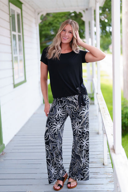 A woman wearing the Coco Palazzo Pants by Rien ne se Perd in Black and White floral, with a wide elastic waistband and wide legs. She is wearing them with a black top and standing on a wooden porch.